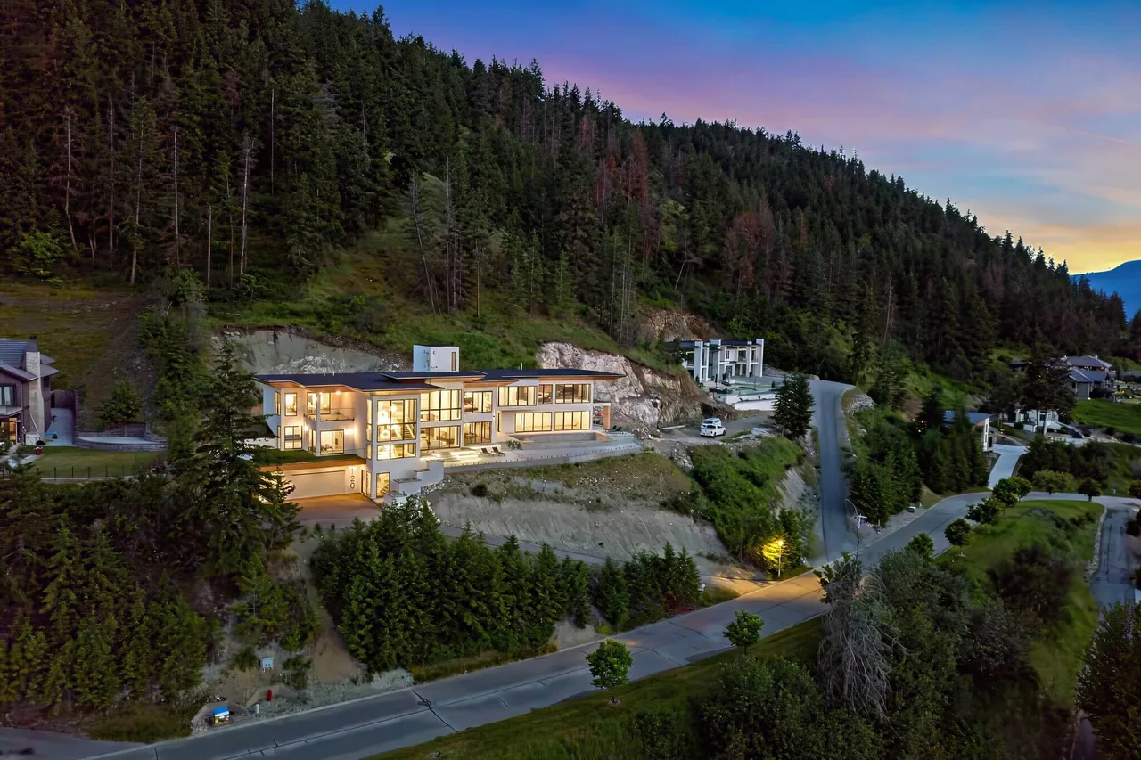 An aerial view at dusk of a lit hillside home among the pines above a winding road