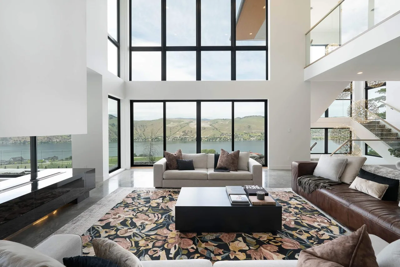 A double-height living room with a patterned rug, leather and white sofas and tall windows onto the lake