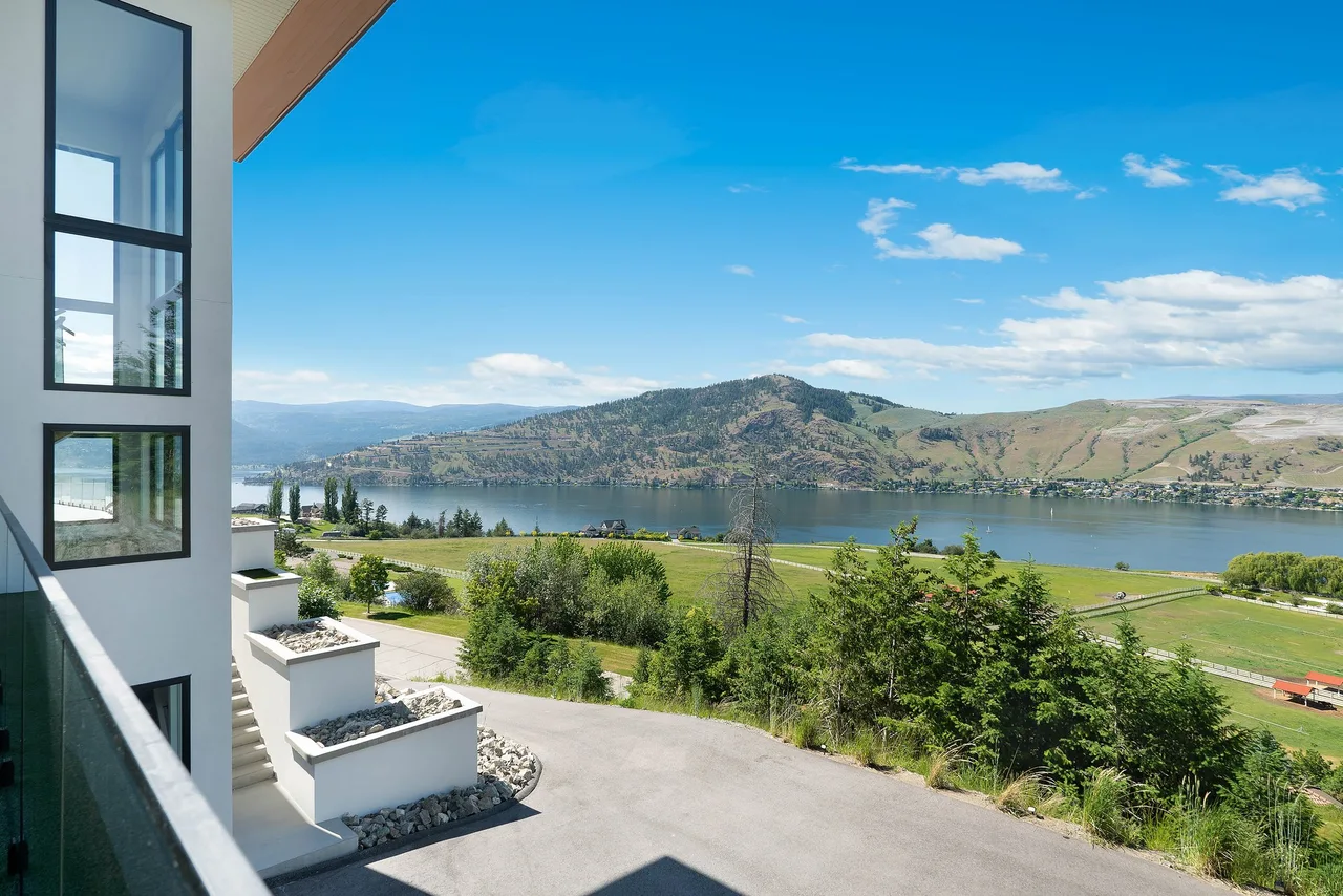 A view from a home’s upper deck over its driveway, green fields and Okanagan Lake